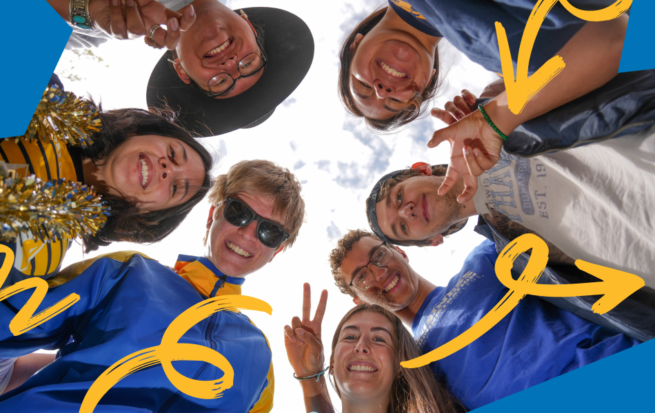 Group of Fort Lewis College students standing in a circle outdoors and looking down toward the camera, with campus buildings and a partly cloudy sky visible above, representing student connection and community.